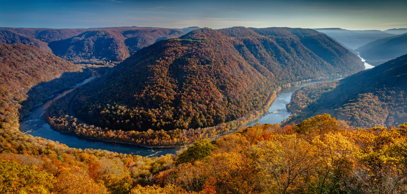 A scenic view of New River Gorge National Park, showcasing a river bordered by lush trees and towering mountains.