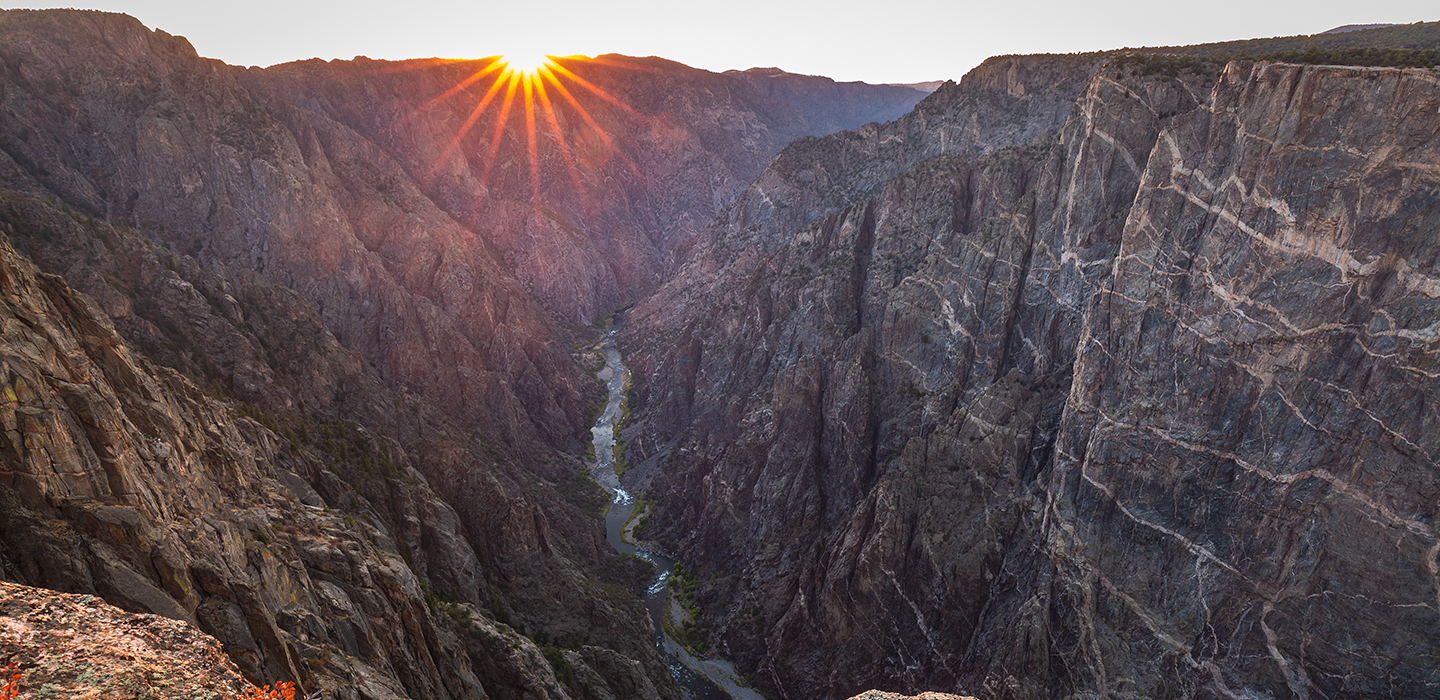The sun brightly shines into a thin river at the base of two mountains at Black Canyon of the Gunnison National Park