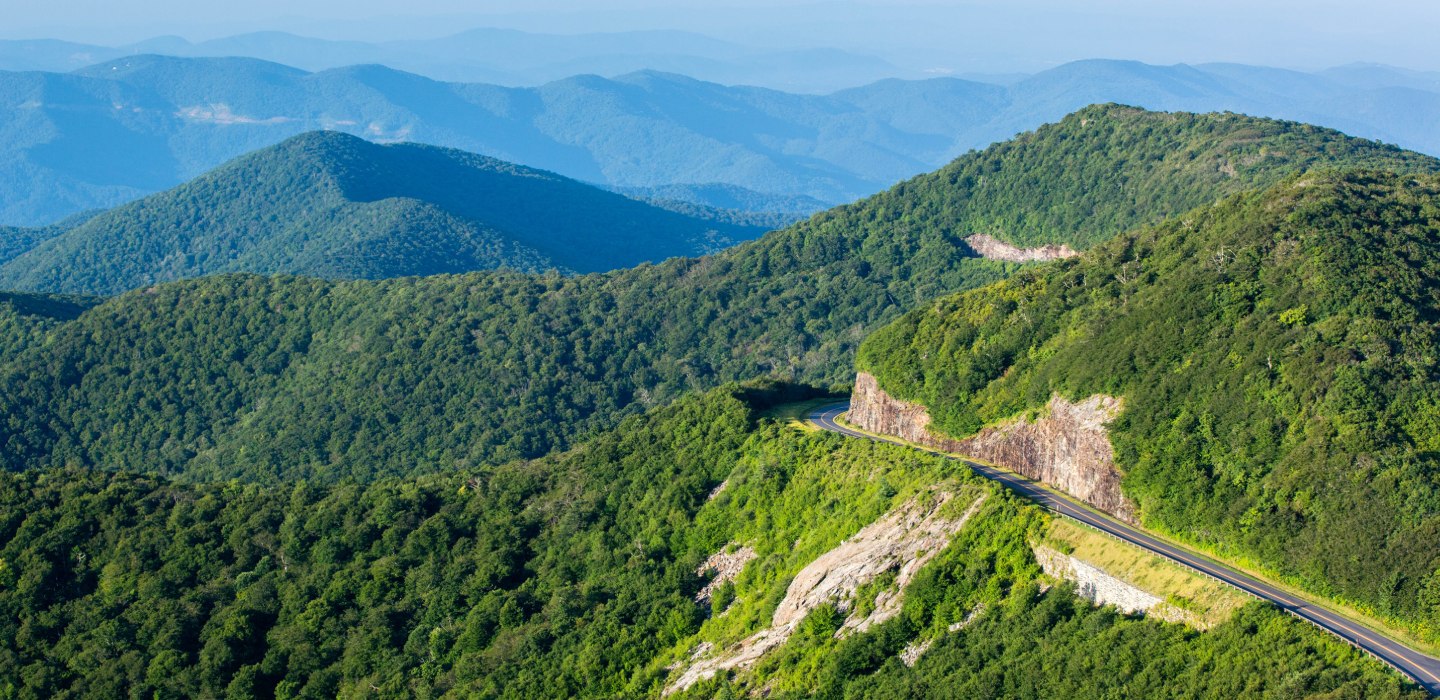 A road winds through the Blue Ridge Parkway's mountains dotted with deep green trees with mountains in the distance.