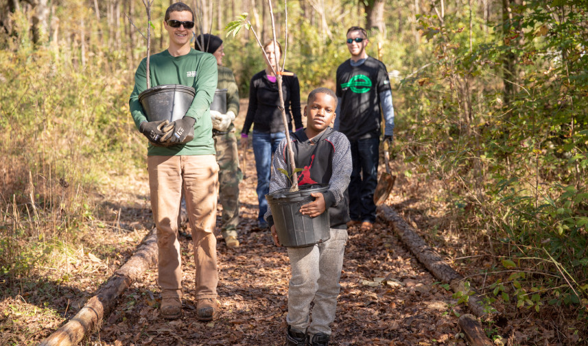 A young boy carries a new growth tree still in its planter along a path with adults behind him