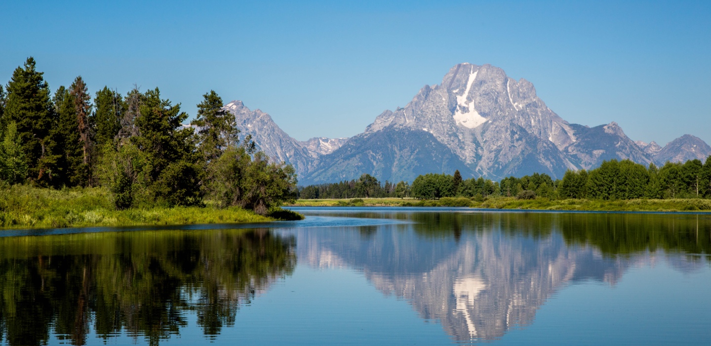 The Grand Tetons reflect in a clear lake with green trees at the Grand Tetons National Park
