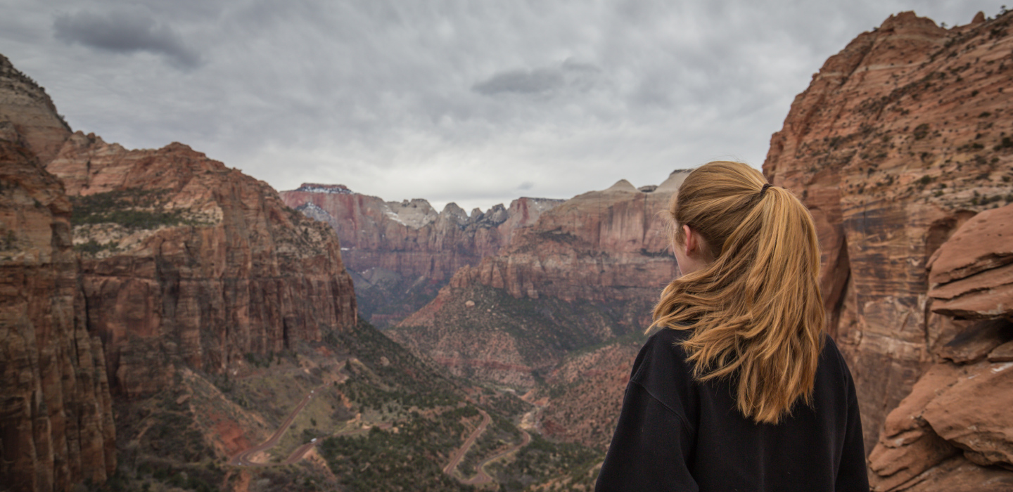 A girl with her hair in a ponytail looks out across Zion National Park on a cloudy day