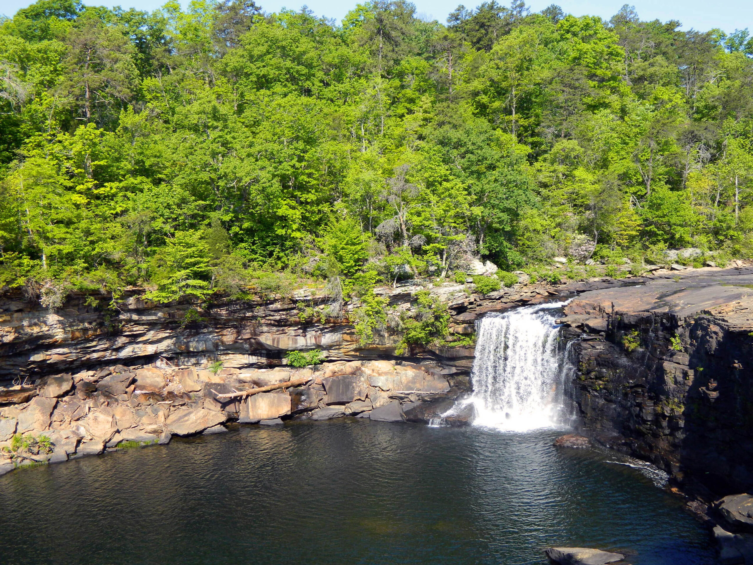 Green trees decorate a small waterfall which flows into a body of water at Little River Canyon