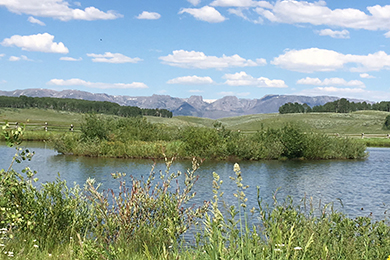 At Loomis Park ranch, a small lake in the foreground is clear and blue. The snowy mountains and tall grasslands are high in the background.