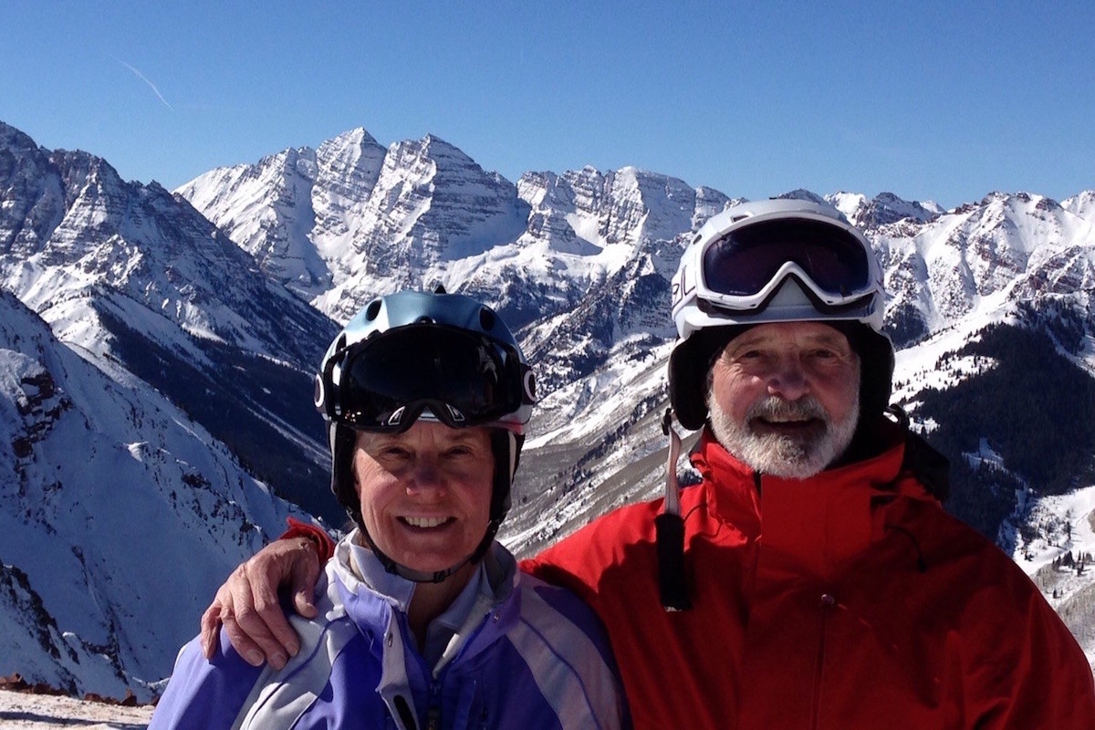 Two people with ski goggles and helmets smiling for the camera with snow covered mountains in the background