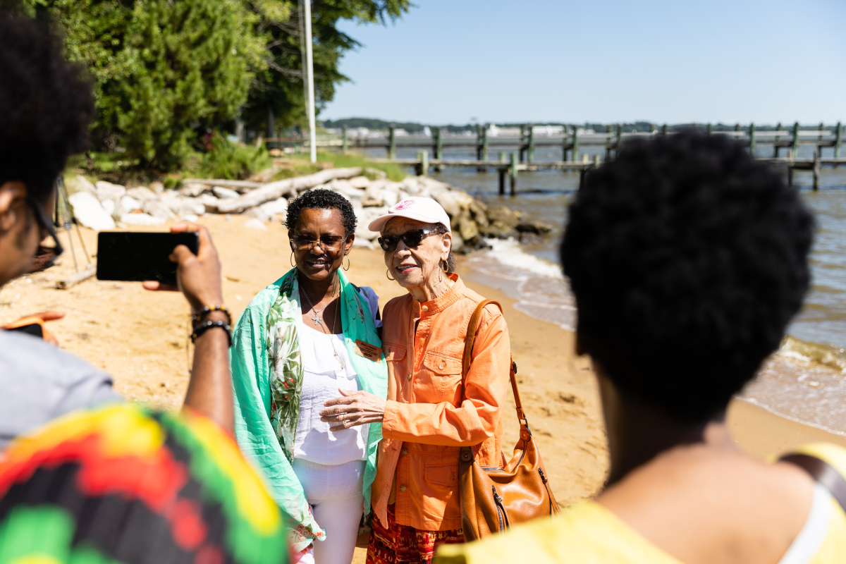 Two women on a beach smiling as two other women look at them, one of them taking a photo