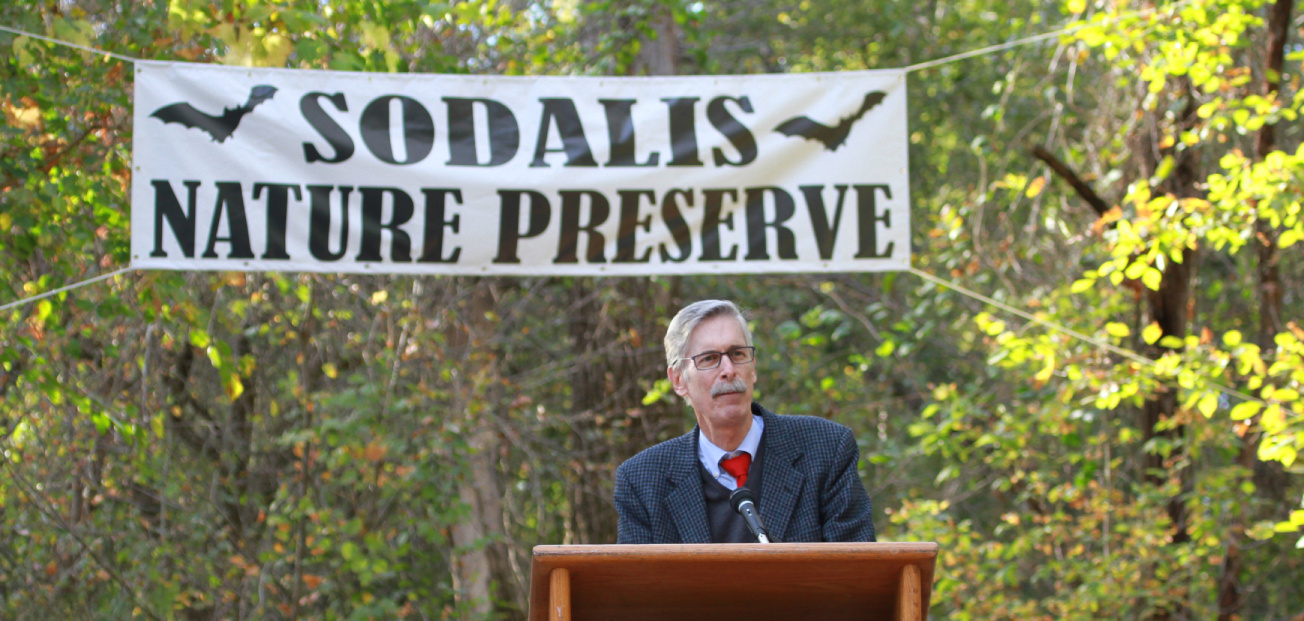 A man stands at a podium, speaking at the opening ceremony of Sodalis Nature Reserve in Hannibal, MO, with a sign behind him.