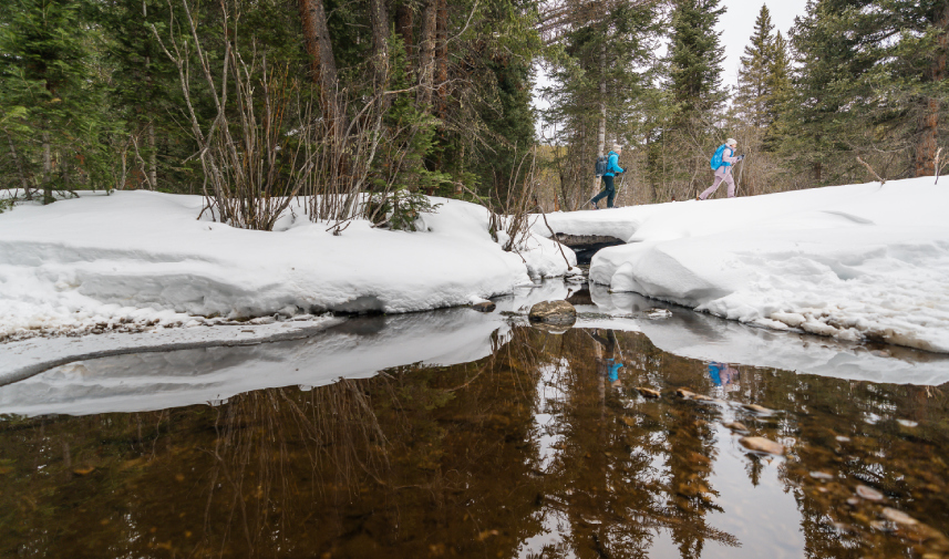 Two people with backpacks walk along a snowy bank beside a creek in Tolland Ranch, with trees and snow reflected in the water.