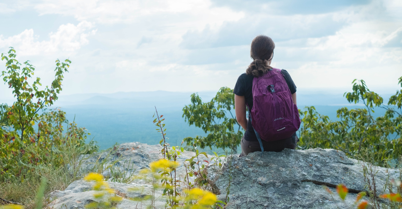 A woman with a backpack sits on a rock, gazing at the mountains in Talladega National Forest.